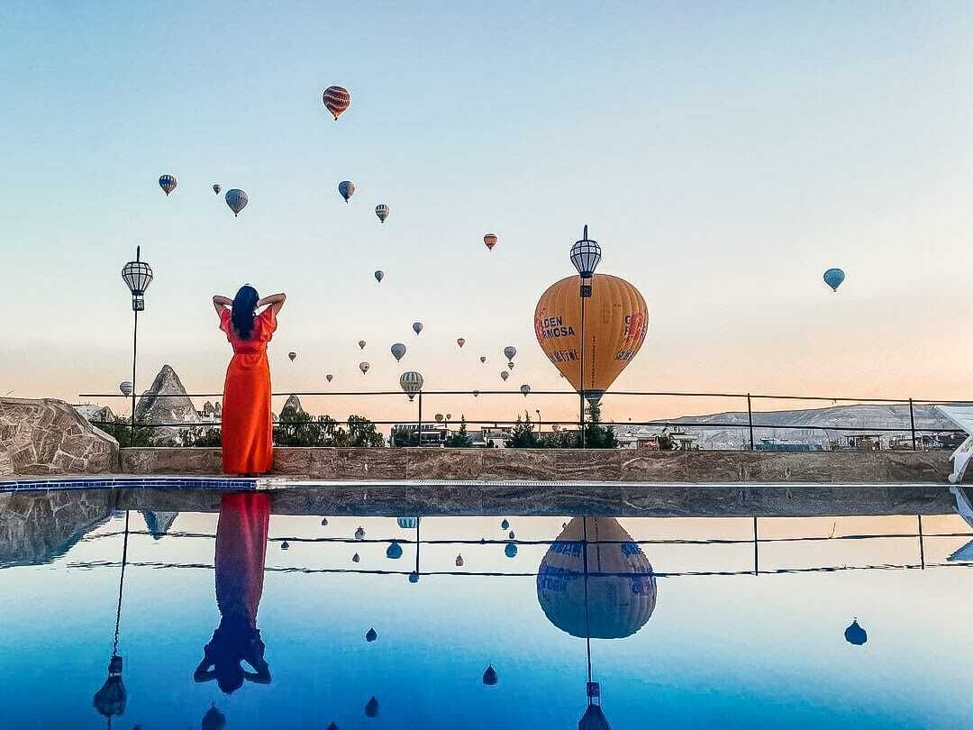 A person in a red dress standing by a reflective pool with hot air balloons in the background.