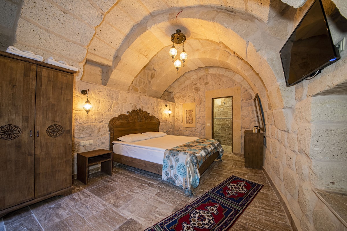 A stone-walled bedroom with a wooden bed, a bench, and a TV mounted on the wall.