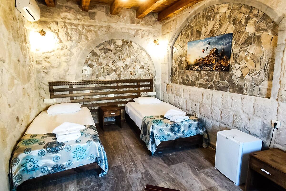 A rustic room with two beds, a wooden bench, a stone wall, and a small refrigerator.