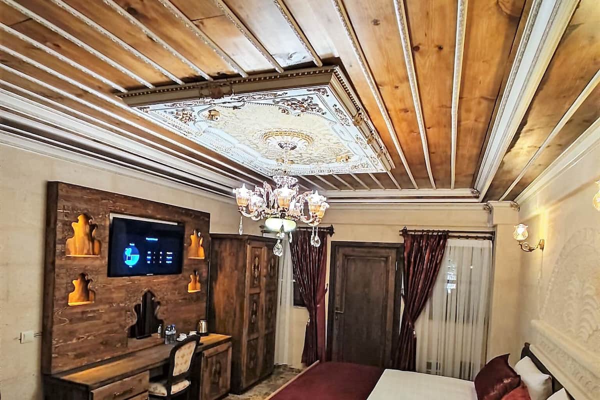 A bedroom with a large bed, wooden ceiling, chandelier, TV, and a desk with a chair.