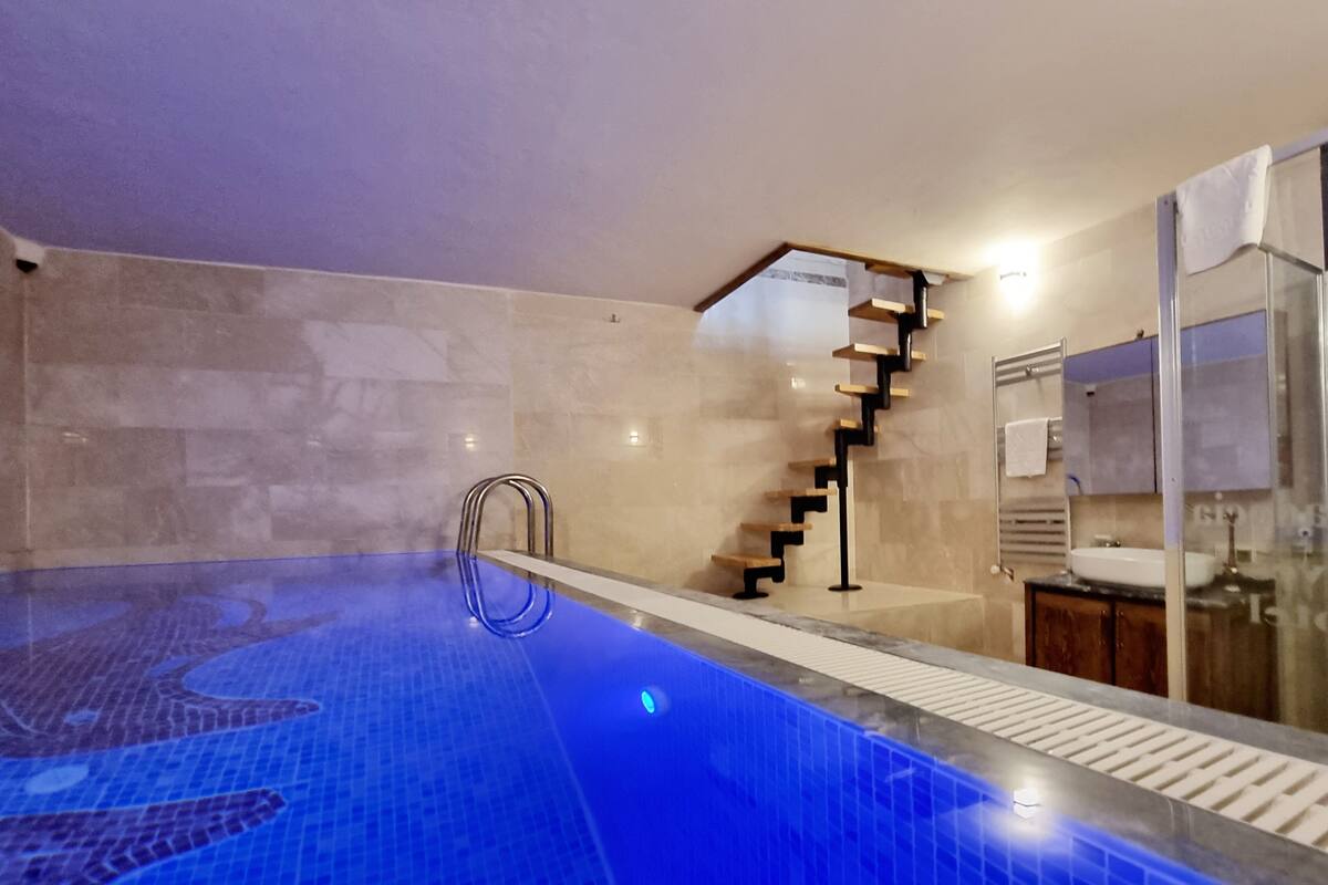 A modern indoor swimming pool with a blue tiled floor, a wooden staircase, and a glass shower enclosure.