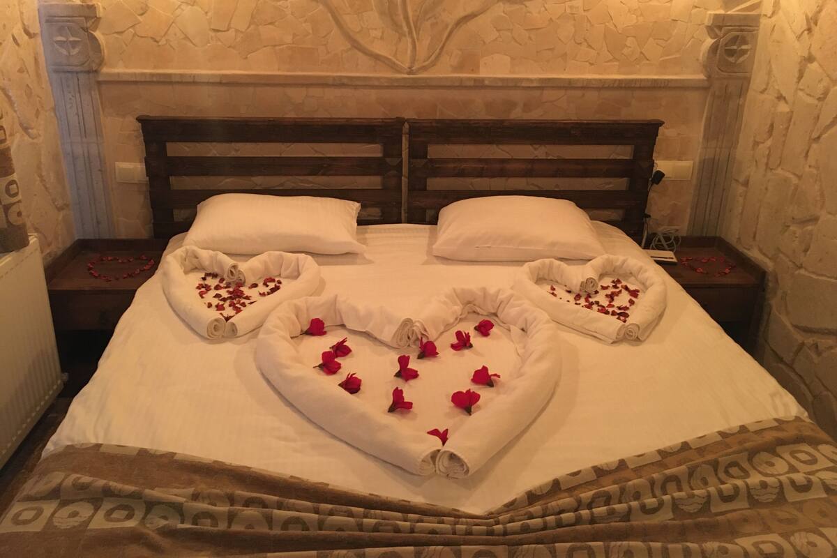 A bed with white linens, a heart-shaped arrangement of rose petals, and a wooden headboard.