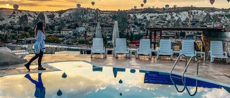 Una piscina con un reflejo nítido del cielo y las nubes, rodeada de sillas de descanso y vistas a un pueblo con edificios y una mezquita.
