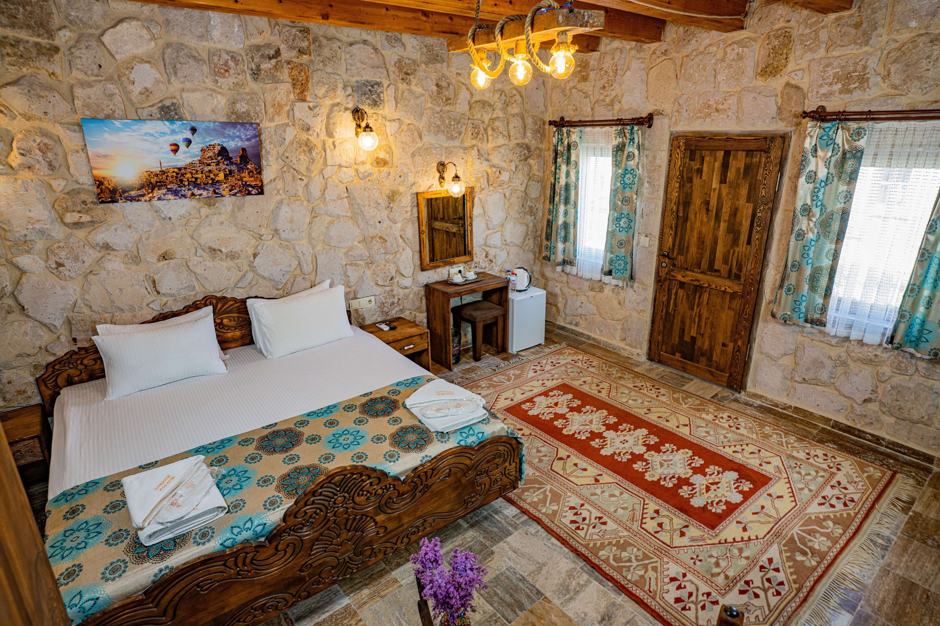 Una camera da letto con parete in pietra, un letto con copripiumino decorato, una testiera in legno, un comodino, una scrivania in legno, una sedia, uno specchio, una porta in legno e una finestra con tende.