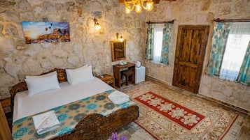 Una camera da letto con parete in pietra, un letto con copripiumino decorato, una testiera in legno, un comodino, una scrivania in legno, una sedia, uno specchio, una porta in legno e una finestra con tende.
