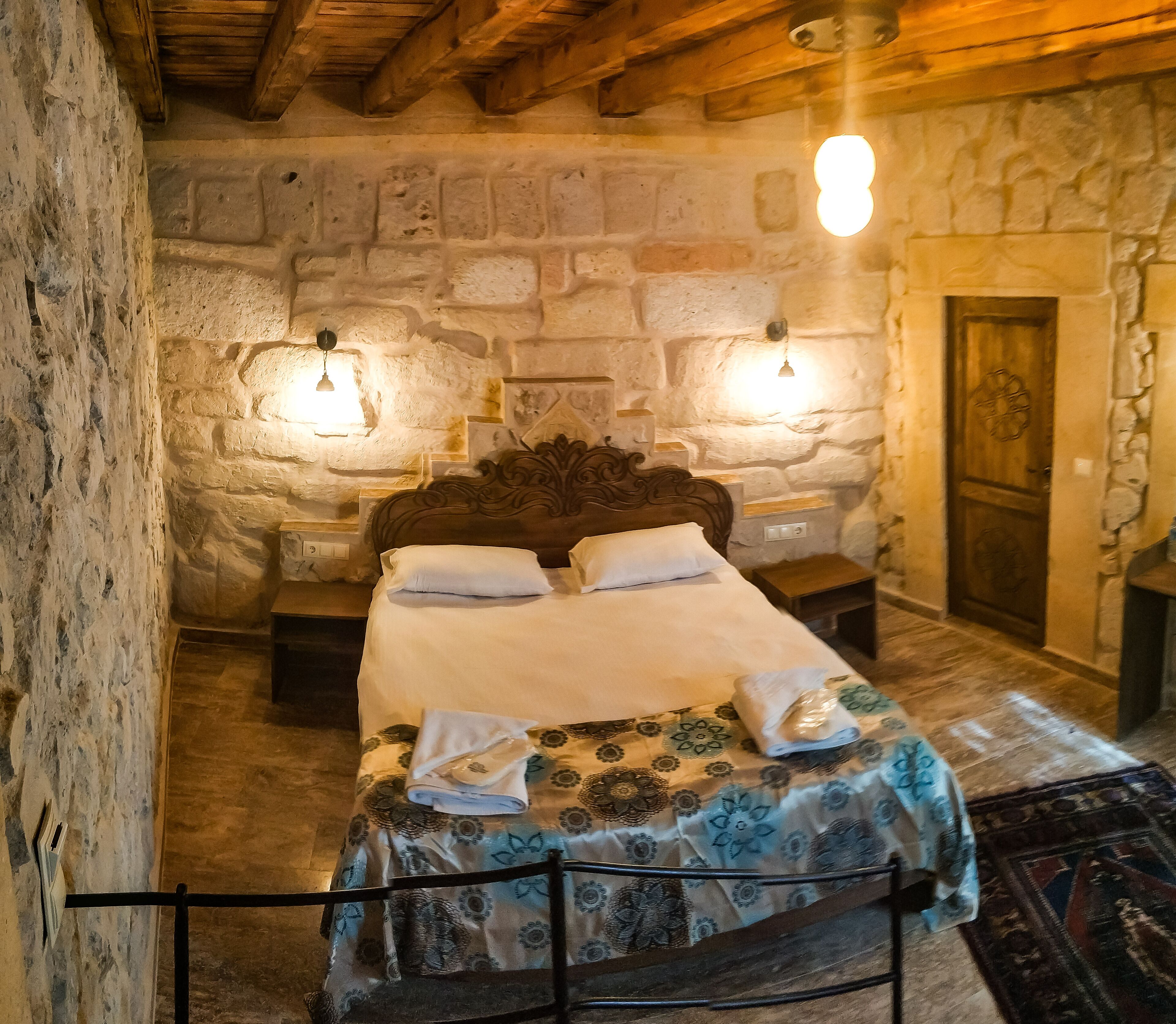 Une chambre aux murs de pierre, avec un lit en bois, deux lampes de chevet et une porte ornée d’un motif circulaire.