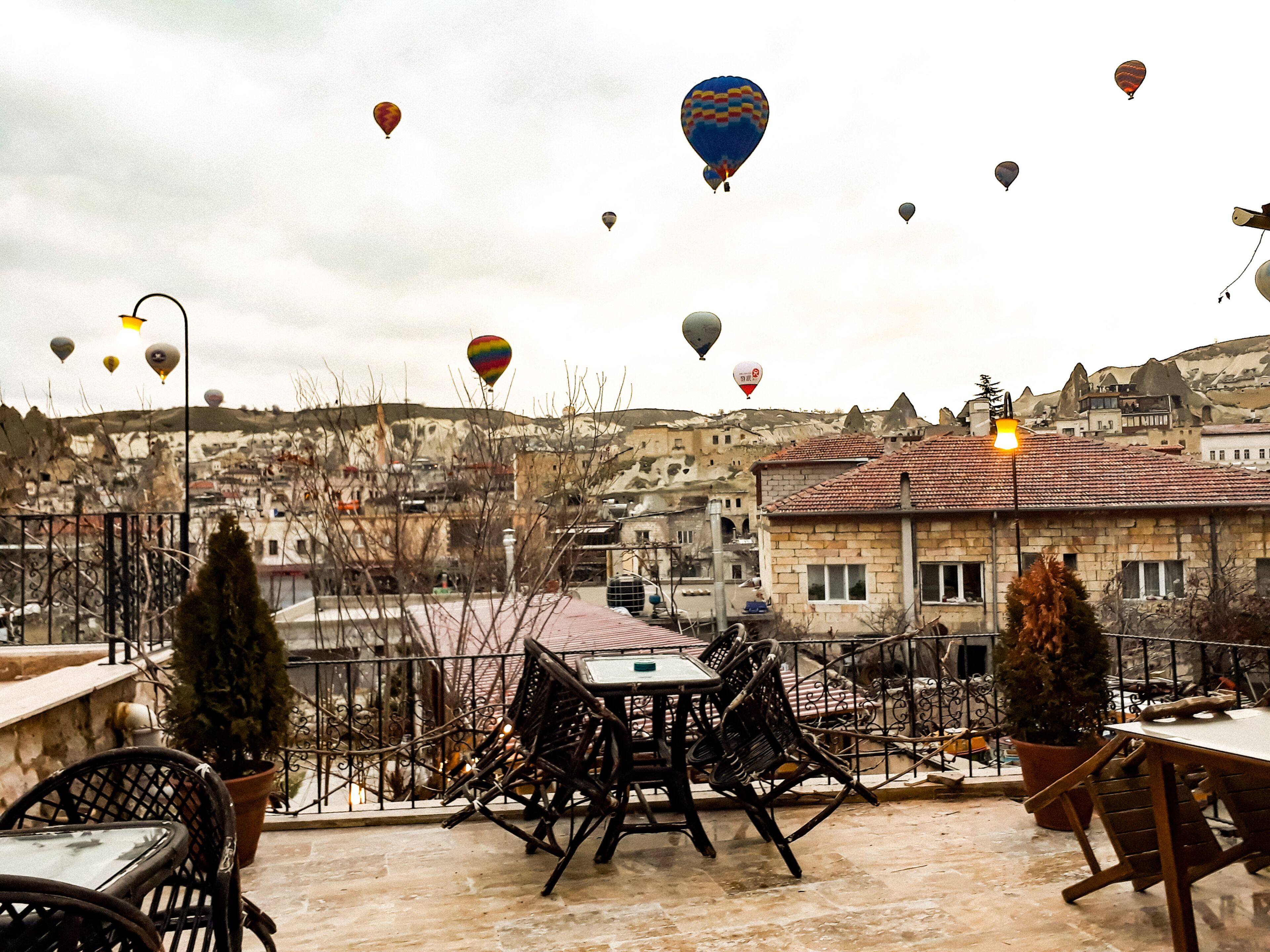 A terrace with outdoor seating, a view of a town with traditional architecture, and hot air balloons in the sky.