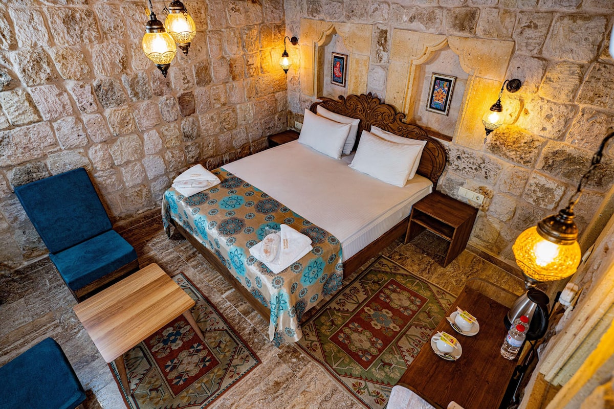 A stone-walled bedroom with a bed, bedside tables, a chair, and a table with a lamp.