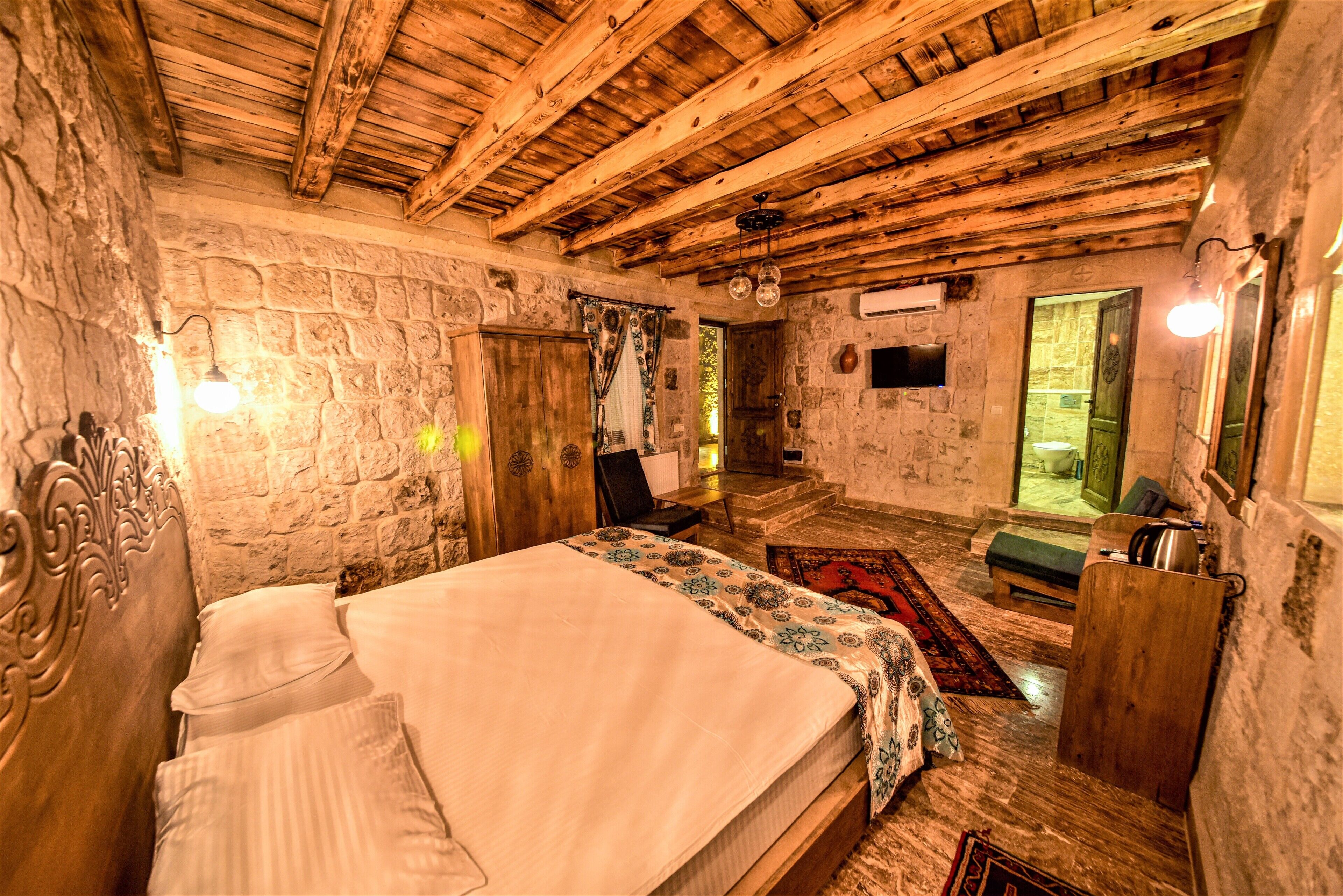 A rustic bedroom with a stone wall, wooden ceiling beams, a bed with a patterned bedspread, a wooden bench, and a small table with a lamp.
