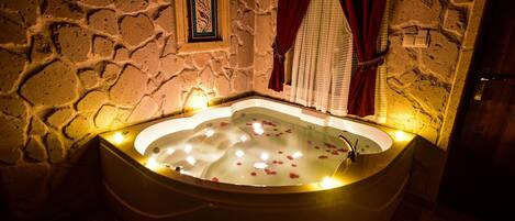 Baño de paredes de piedra con una bañera independiente llena de agua y pétalos de rosa, iluminado por una luz cálida.