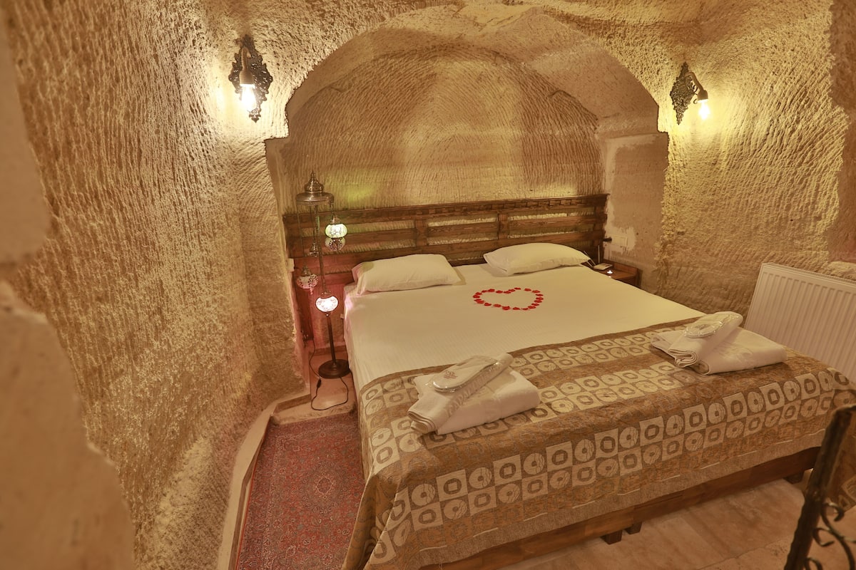 A room with a bed, a wooden headboard, a nightstand with a lamp, and a wall-mounted light fixture.