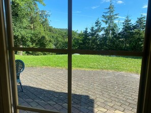 A view through a window with a brick patio, green lawn, and trees. - Double room (apartment 2) - log cabin (Sangerhausen)