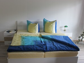 Una cama bien hecha con una colcha azul y amarilla, dos almohadas, una mesita de noche con una lámpara y un aparador con un libro.