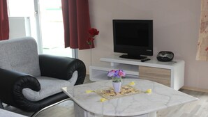 Ein modernes Wohnzimmer mit einem grauen Sofa, einem weißen Couchtisch mit Blumenvase, einem Flachbildfernseher auf einer weißen Konsole und einem roten Vorhang.