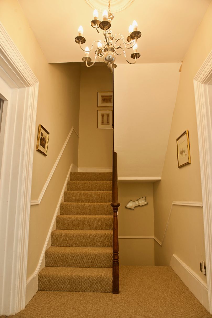 A staircase with carpeted steps and a wooden handrail, leading to a landing with a chandelier and framed pictures on the walls.