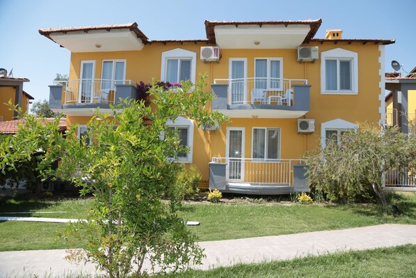 A yellow two-story building with balconies, air conditioning units, and a paved pathway leading to the entrance.