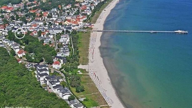 Vista aérea de un pueblo costero con un muelle largo que se extiende hacia el mar, rodeado de edificios residenciales y una playa.