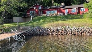 A red building with a dock extending into a pond, surrounded by trees. - Private on Chain O’Lakes! Huge Kitchen, 2 Living Spaces, Large Cousins Room! (Lac Du Flambeau)
