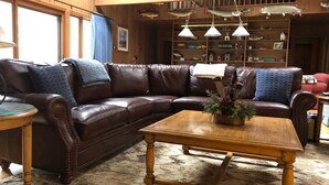 A cozy living room with a brown leather sectional sofa, a wooden coffee table, and a bookshelf filled with various items. - Private on Chain O’Lakes! Huge Kitchen, 2 Living Spaces, Large Cousins Room! (Lac Du Flambeau)
