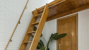 Una escalera de madera apoyada en una pared de ladrillo blanco, una puerta de madera y una planta en maceta.