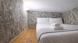 Un dormitorio con cama, ropa de cama blanca, suelo de madera y papel tapiz floral.