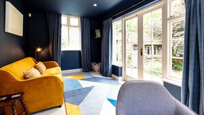 Een kamer met een gele bank, een grijze stoel en een kamerbreed tapijt met een patroon. Er zijn grote ramen met gordijnen, een deur en een wandlamp.