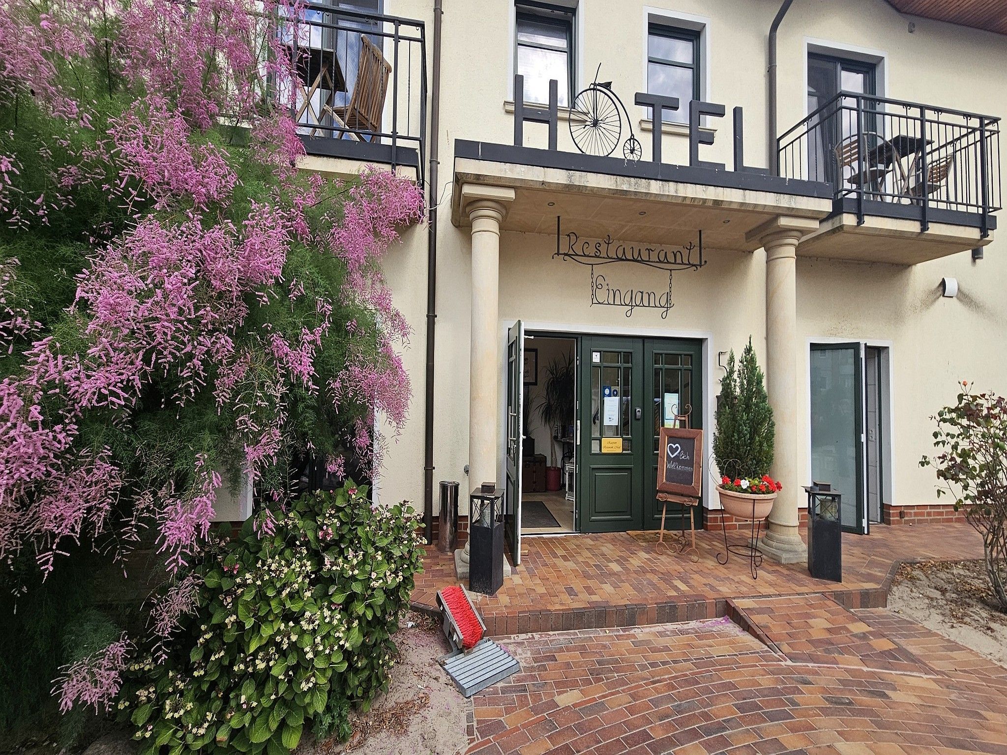 Um restaurante com varanda, uma placa sobre a entrada e um vaso de flores no chão.
