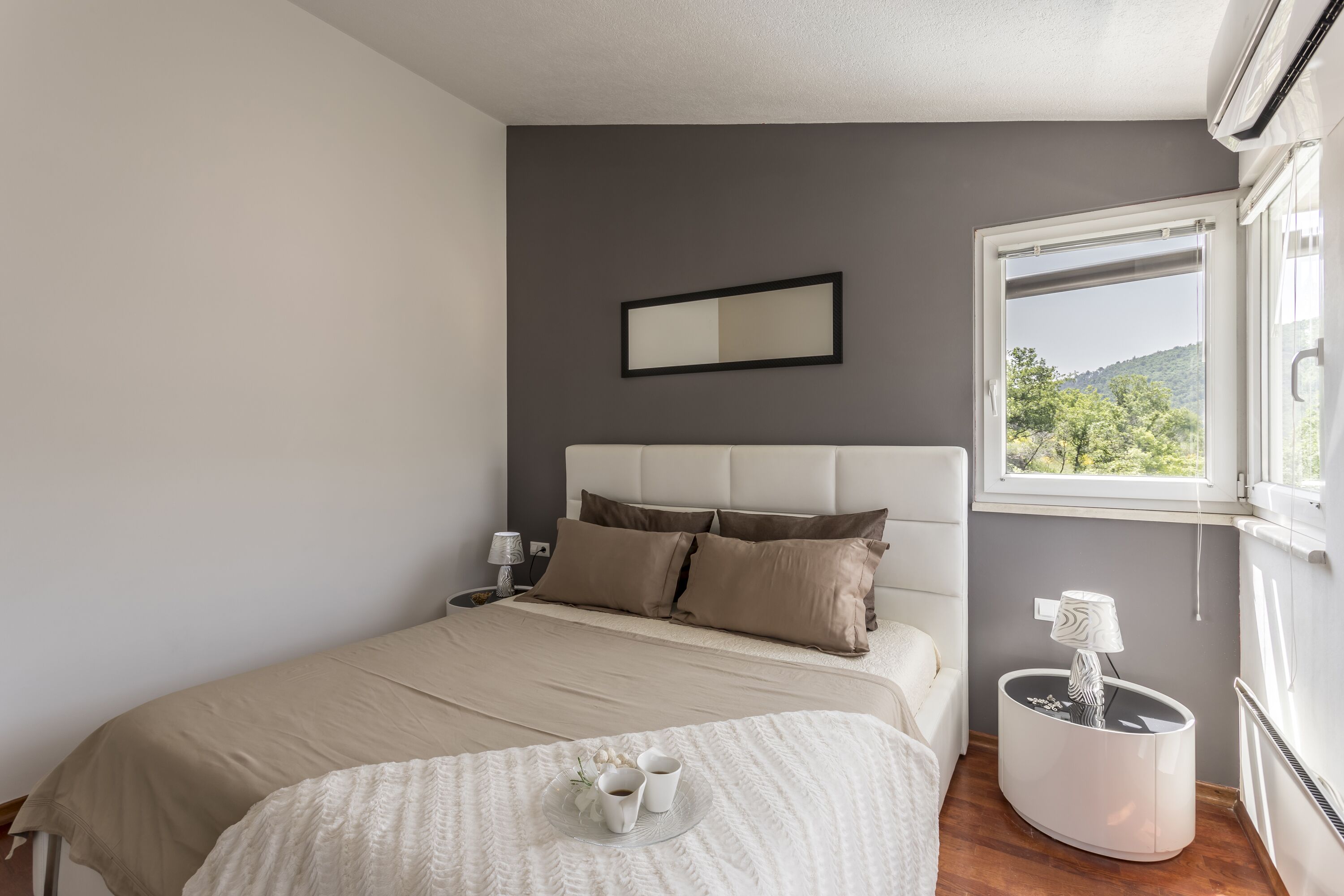 A bedroom with a bed, bedside table, lamp, and a view of greenery outside.