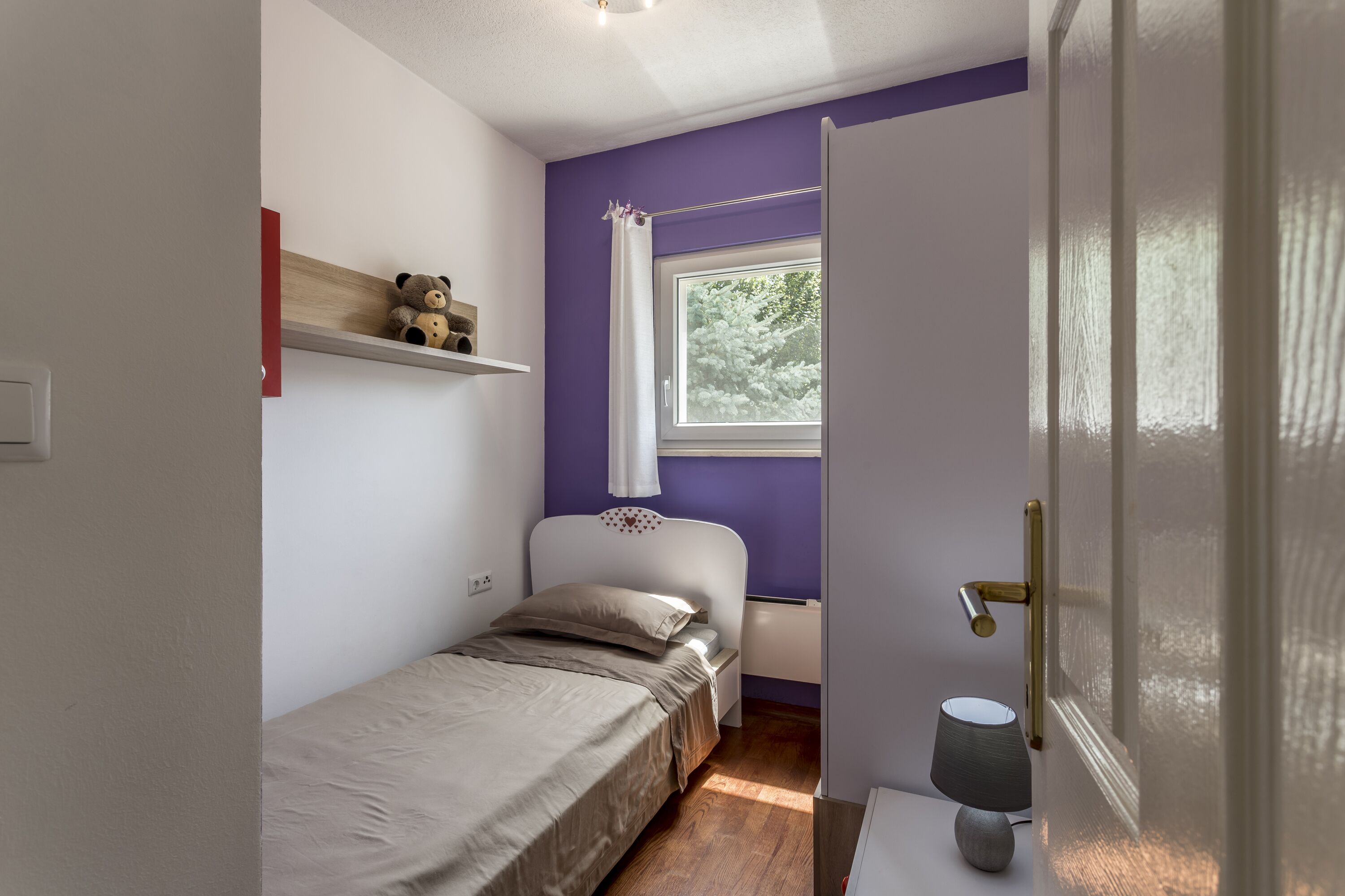 A small, neatly organized bedroom with a single bed, a nightstand, a lamp, and a window with a view of trees.