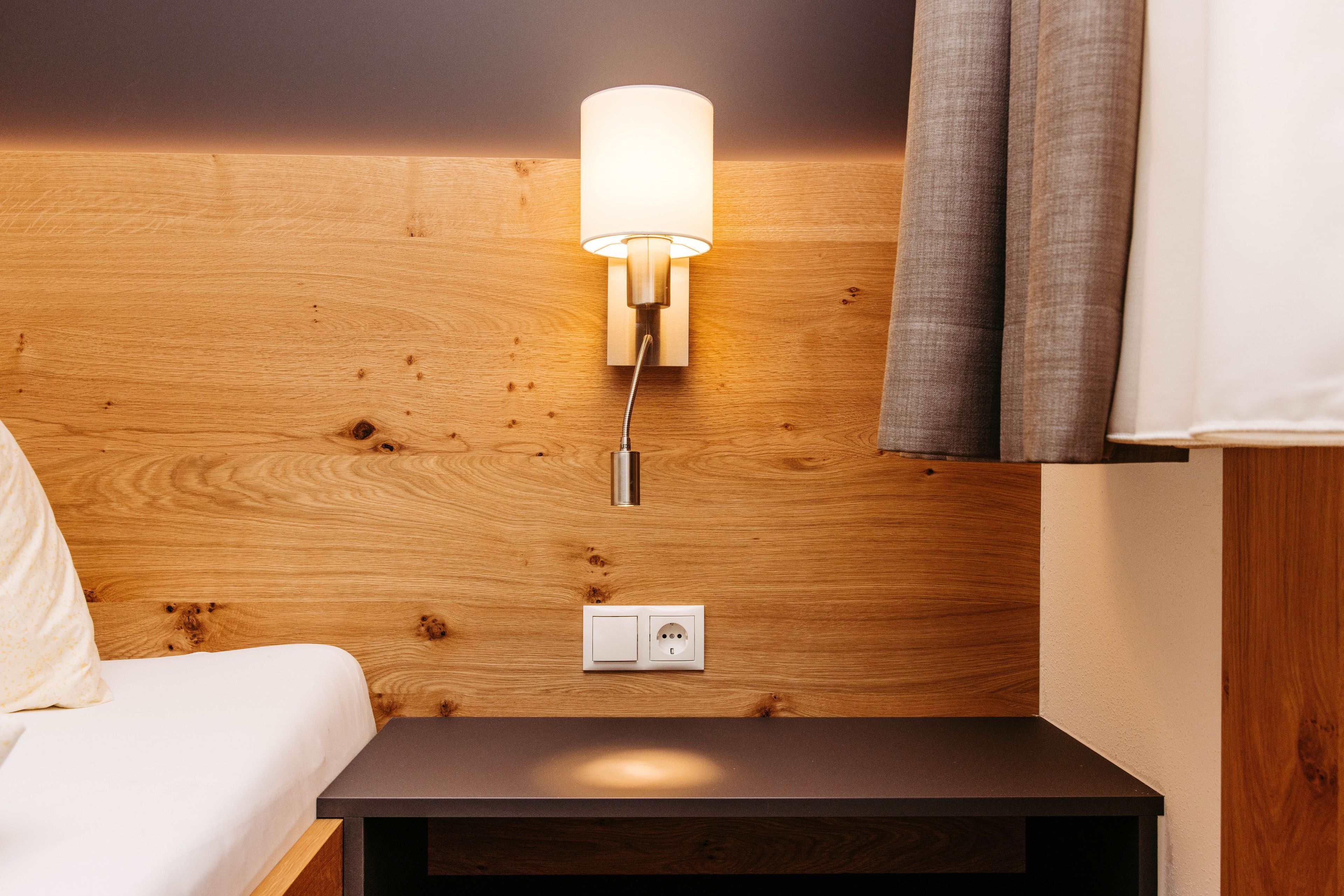 Une tête de lit en bois, un luminaire fixé au mur, une prise de courant et une table de chevet.