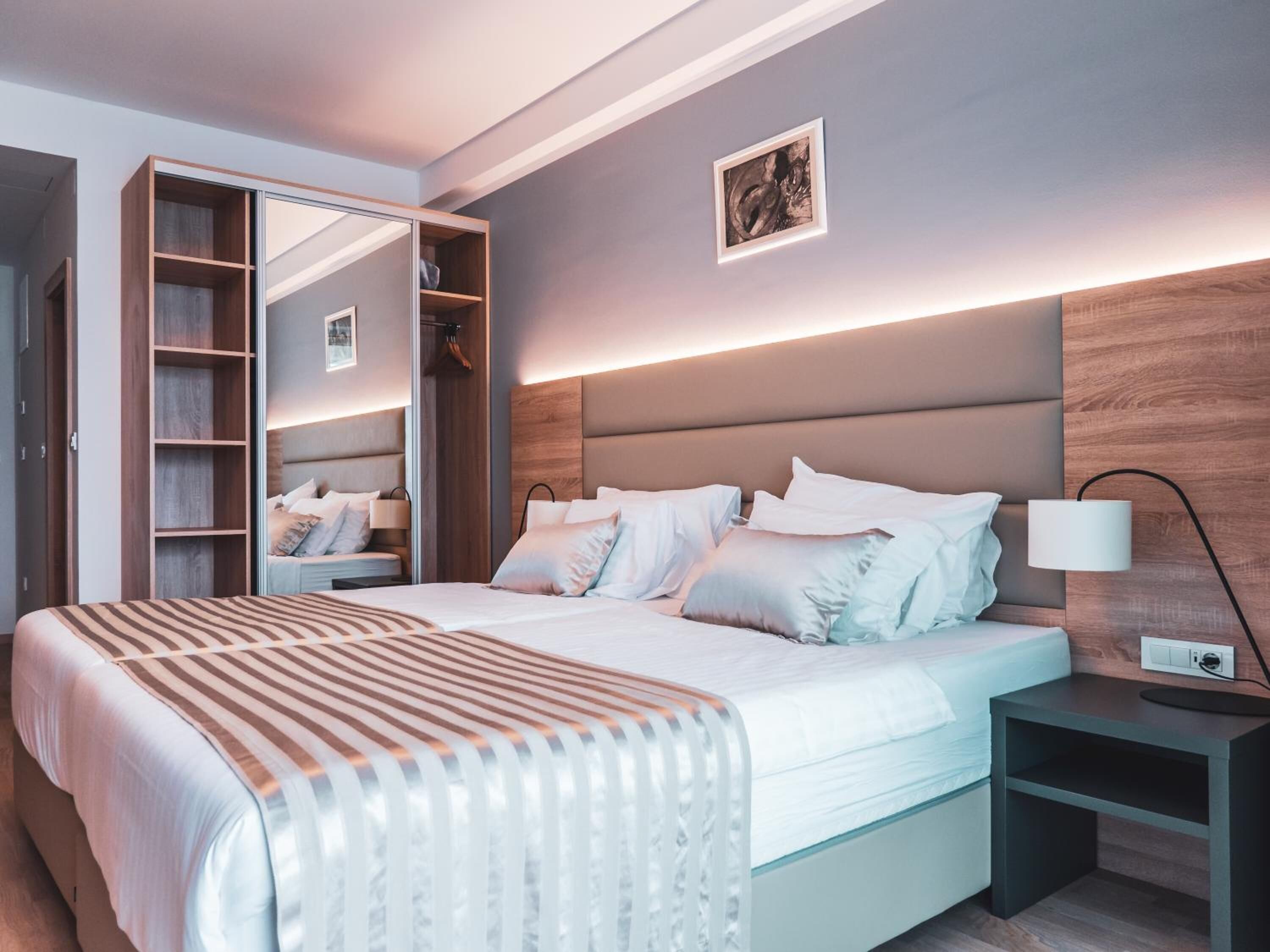 A modern bedroom with a large bed, a bedside table, a nightstand, a wardrobe with a mirror, and a wall-mounted lamp.