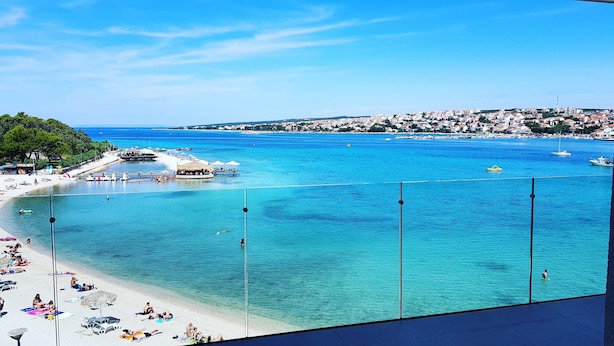 A coastal view with a beach, clear blue waters, and a town in the background. - Hotel Kaneo (Novalja)