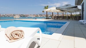 A poolside area with a view of a coastal town, lounge chairs, and umbrellas. - Hotel Kaneo (Novalja)