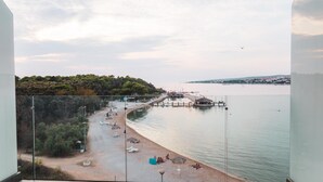 A balcony with a view of a beach, a pier, and a forested hill. - Hotel Kaneo (Novalja)