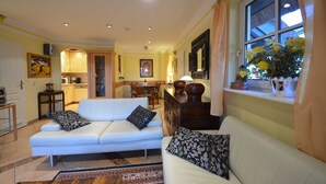 Ein Wohnzimmer mit einem weißen Sofa, gemusterten Kissen, einem Essbereich mit Tisch und Stühlen sowie einem Fenster mit Blick auf Blumen.