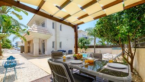 Un espace de restauration en plein air avec une table dressée pour deux personnes, entourée de chaises en osier, sous une pergola.