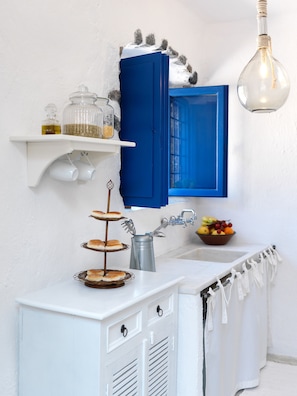 Una kitchenette con encimera blanca, un mueble azul, una lámpara colgante y un bol con fruta.
