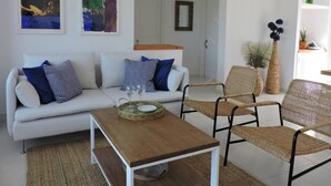 Ein modernes Wohnzimmer mit einem weißen Sofa, zwei Sesseln, einem Holztisch und einem kleinen Beistelltisch. An den Wänden hängen gerahmte Kunstwerke, es gibt einen Spiegel und einen Blumentopf.