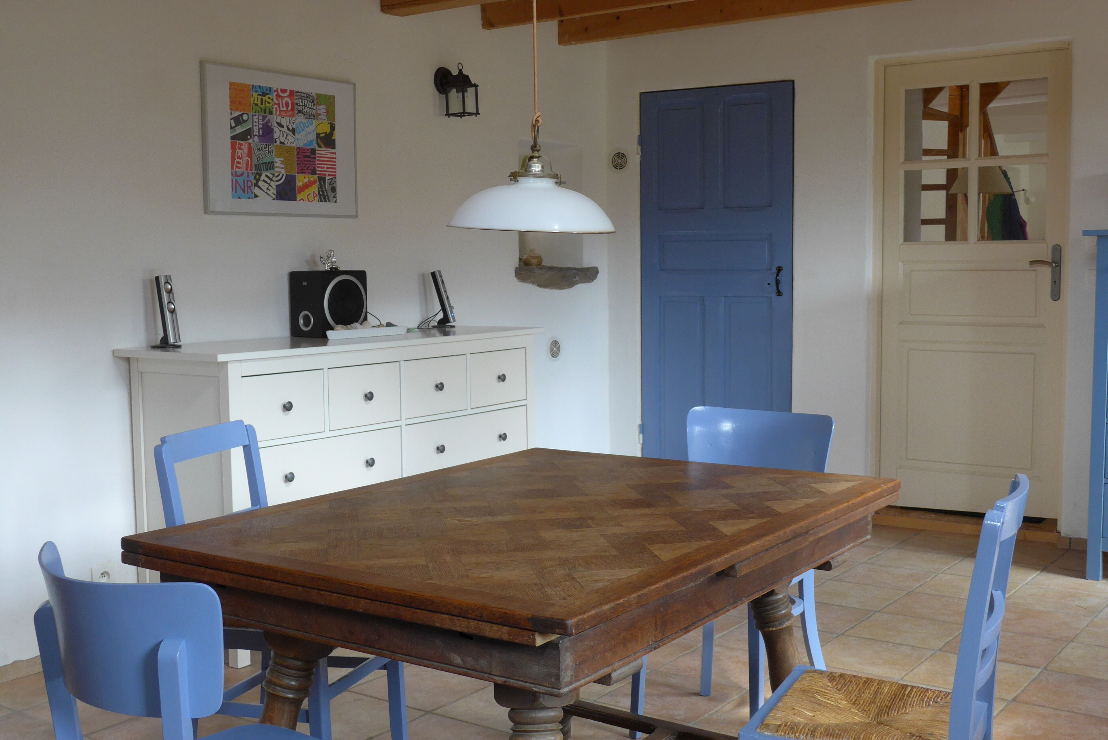 Un coin repas avec une table en bois, des chaises bleues, un meuble blanc avec des tiroirs, une porte bleue et un luminaire suspendu.