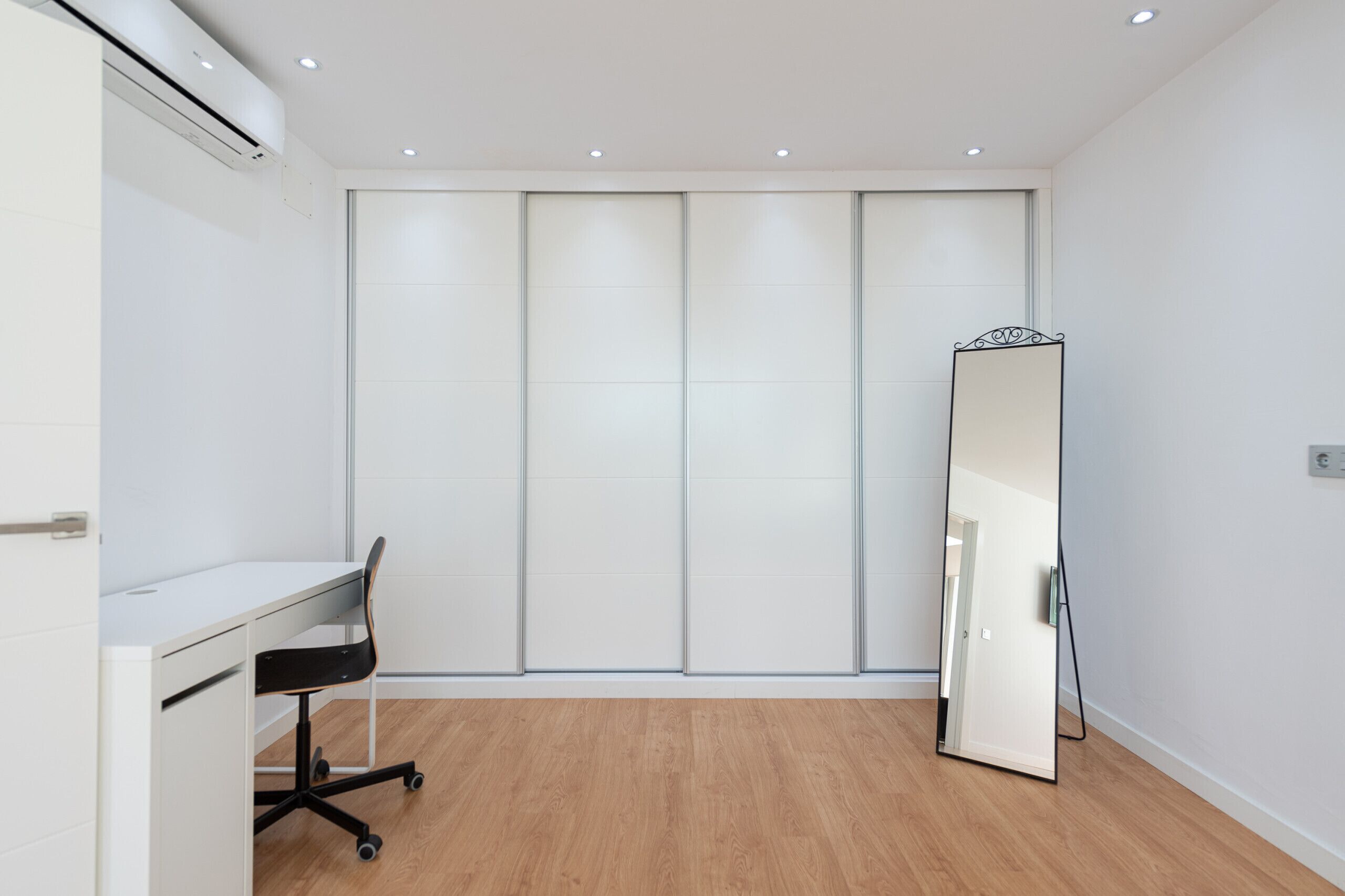 Ein Zimmer mit weißem Schreibtisch, schwarzem Bürostuhl und einem großen Spiegel.