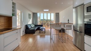 Eine moderne Küche mit Einbauofen und Edelstahlkühlschrank. Ein Essbereich mit Barhocker und einem kleinen Tisch ist vorhanden. Das Fenster bietet einen Ausblick ins Freie.