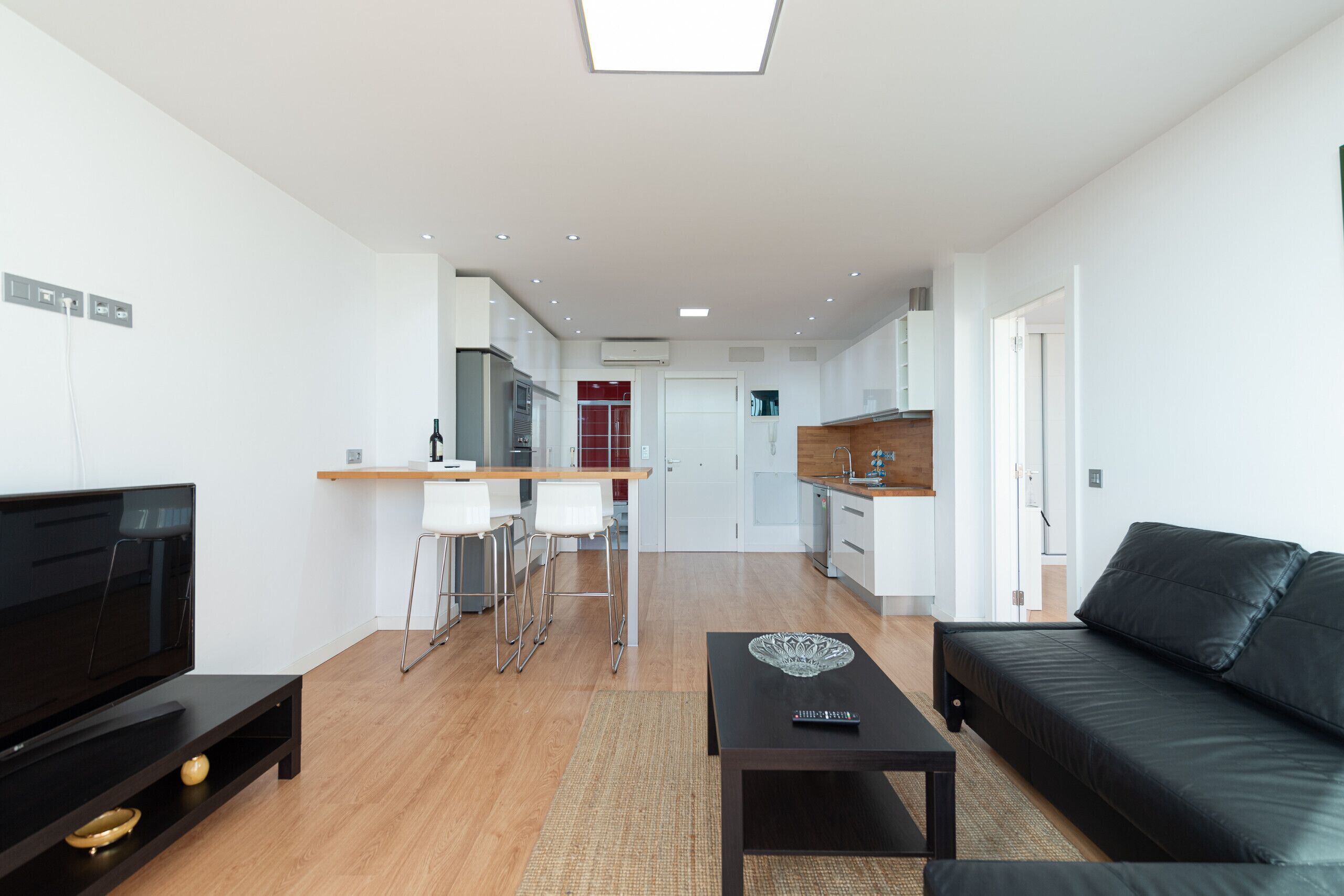 Un appartement moderne comprenant un salon, une cuisine et un coin repas. On y trouve un canapé en cuir noir, un téléviseur et une petite table agrémentée d’un objet décoratif.