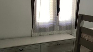 Ein Schlafzimmer mit Bett, Vorhängen am Fenster, einem Nachttisch und einer Kommode mit Schubladen.