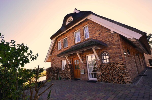 A brick house with a front porch, a wooden door, and a stack of firewood. - 8a Reet-house part Sonnenkliff OG 3 bedrooms - Thatched-roof vacation apartments 8 Kröning (Gager)