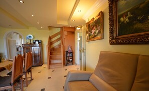 A living room with a sofa, wooden chairs, a staircase, a dining area with a table and chairs, and a painting on the wall.