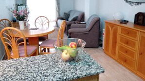 A dining area with a round table, chairs, and a bowl of fruit. There is a sofa and a cabinet in the background.