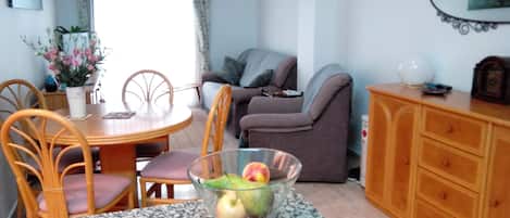 A dining area with a round table, chairs, and a bowl of fruit. There is a sofa and a cabinet in the background.