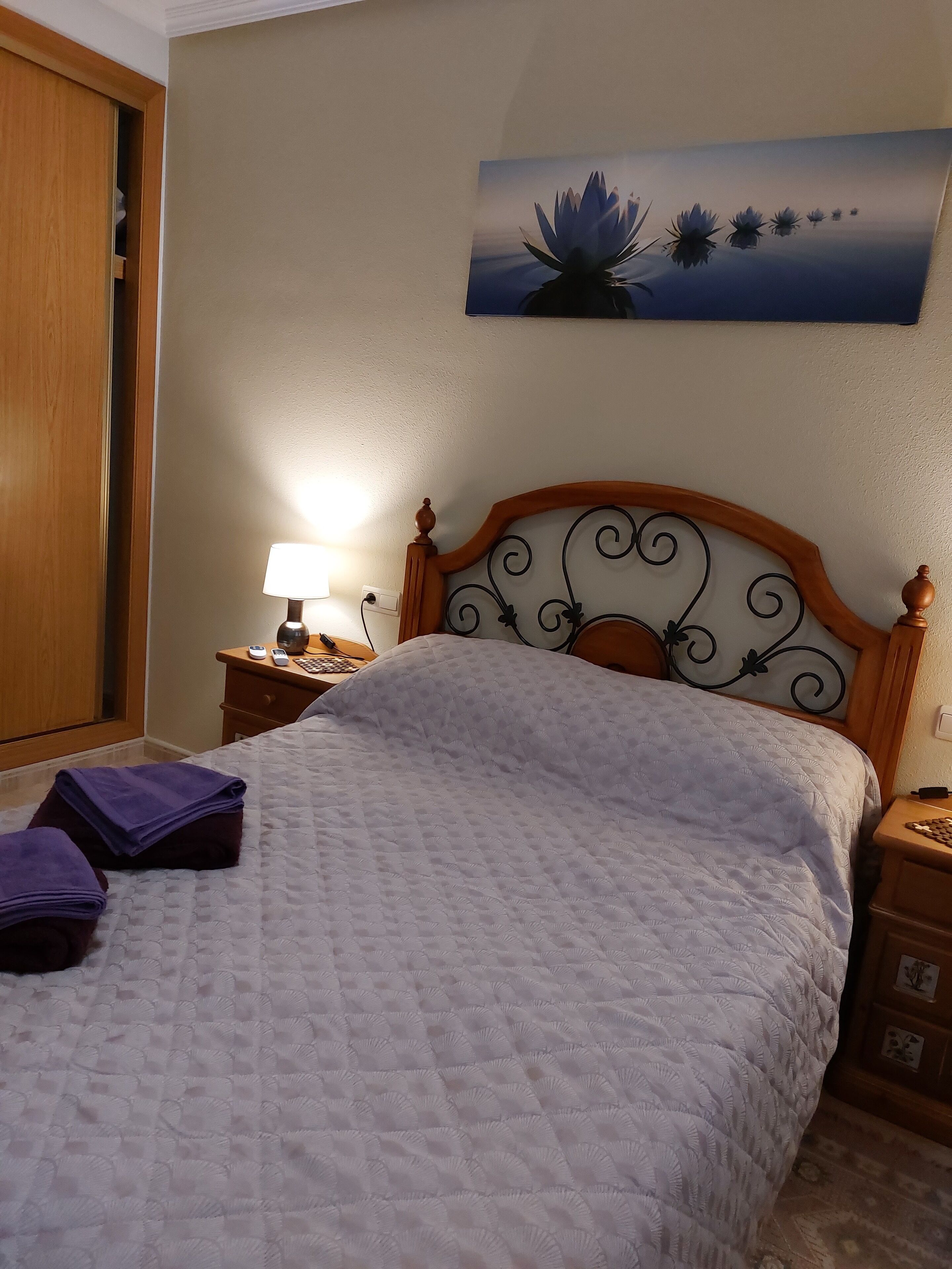 A neatly made bed with a quilted bedspread, a wooden headboard with a decorative design, a bedside table with a lamp, and a wall-mounted artwork of lotus flowers.