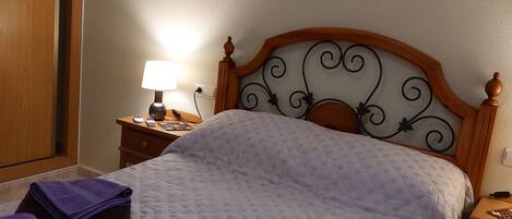 A neatly made bed with a quilted bedspread, a wooden headboard with a decorative design, a bedside table with a lamp, and a wall-mounted artwork of lotus flowers.