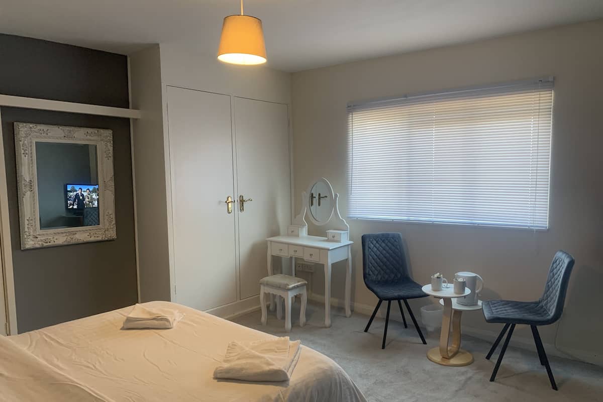A hotel room with a bed, a vanity with a mirror, a chair, a small table with a cup, and a window with blinds.
